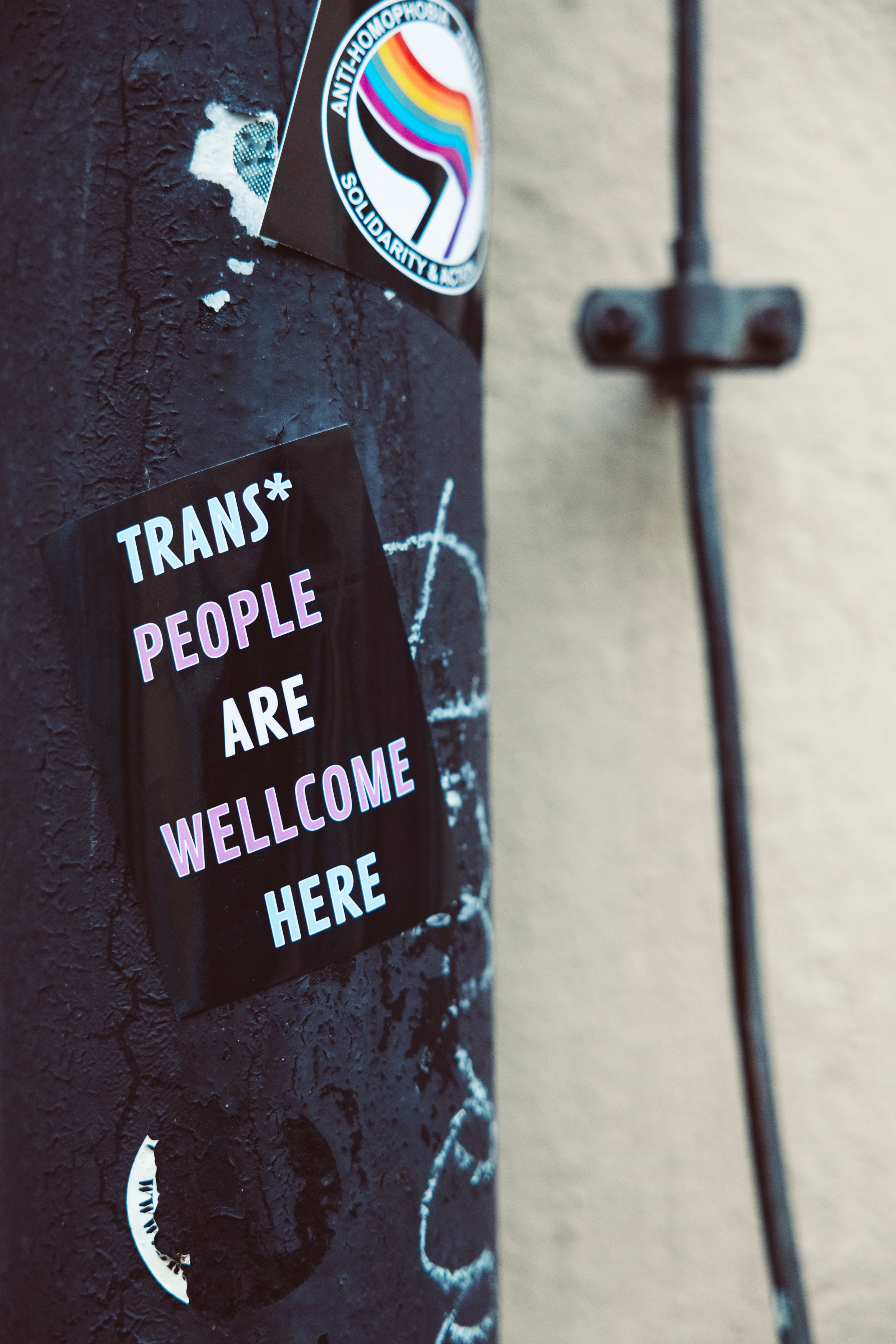 Imagem de um poste com adesivos: um deles em foco escrito 'Trans* people are wellcome here